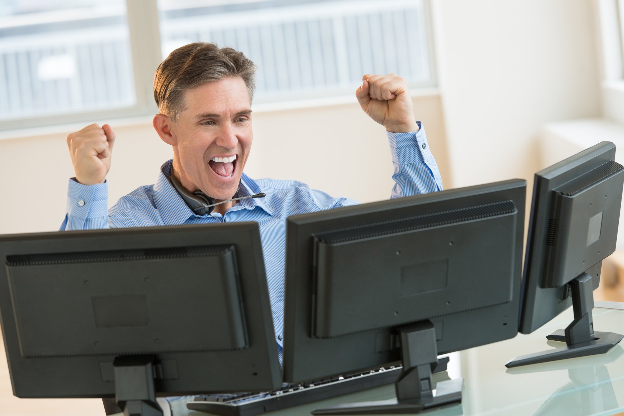 Investor raising his hands in the air with happiness as he looks at three computer screens