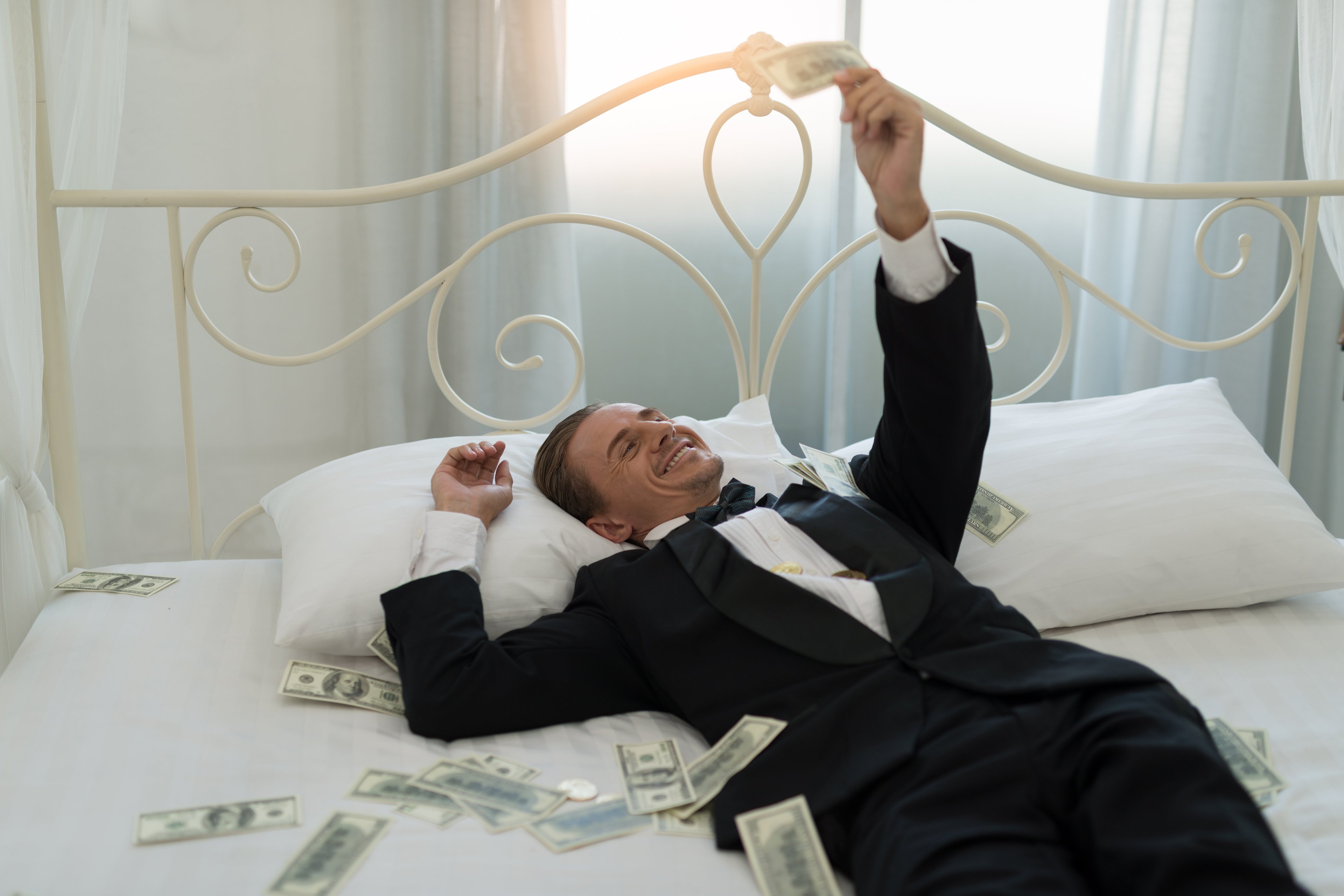 Man in tuxedo lying in a bed with money.