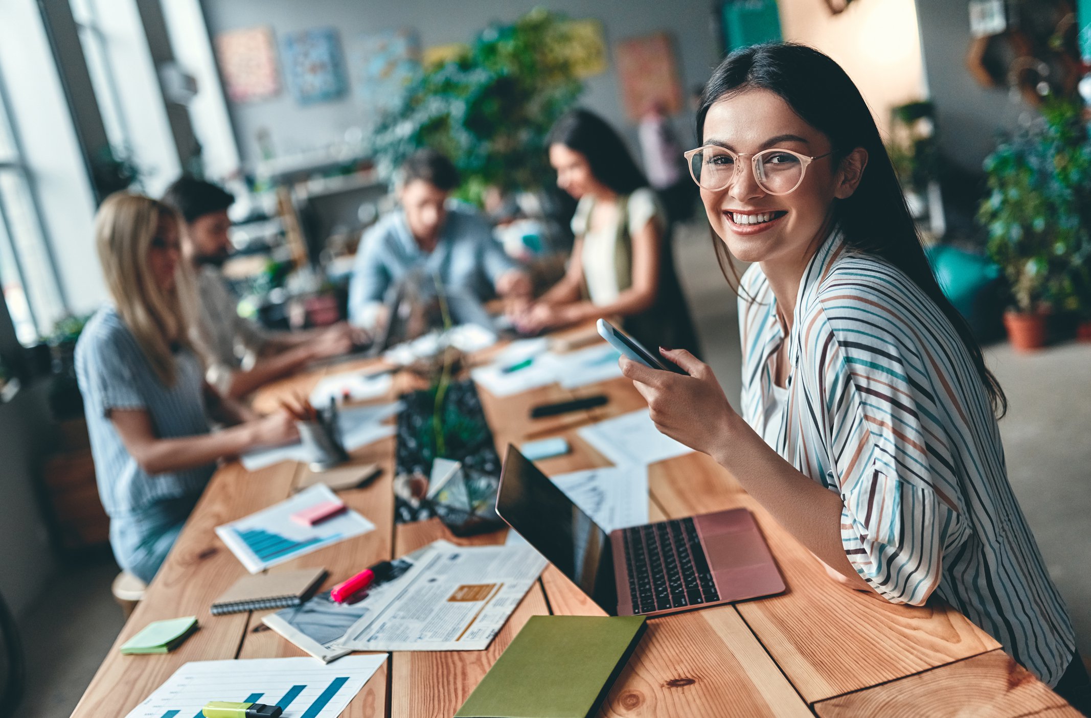 Woman in striped shirt and glasses smiling and sitting at long wooden table with other remote workers