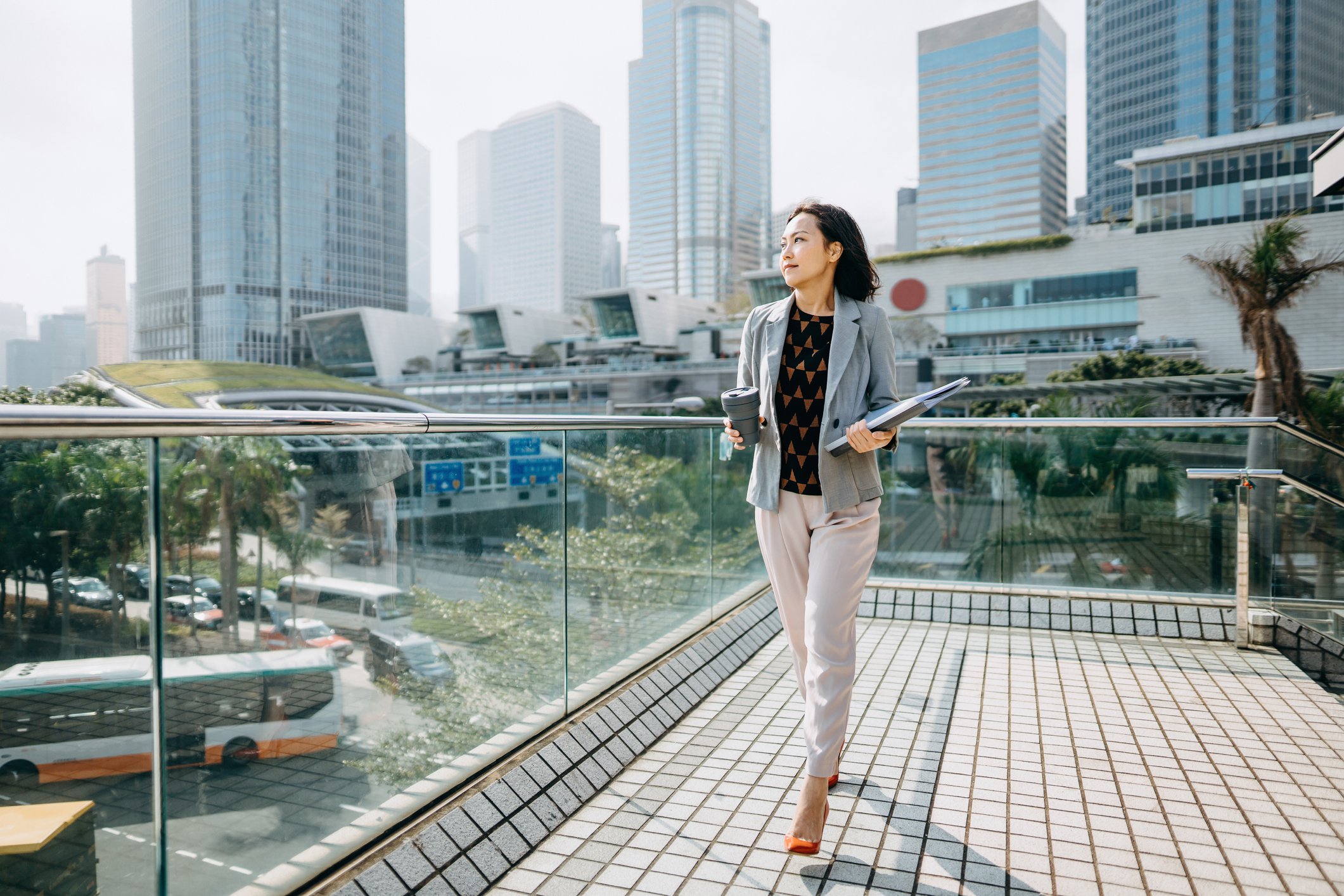 Businessperson walking with folder of papers on rooftop balcony of building and staring out at city filled with skyscrapers