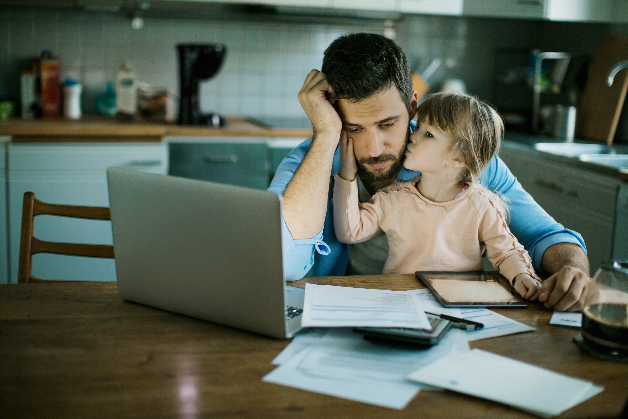 Stressed person looking at laptop and documents with young child sitting on lap kissing their face.