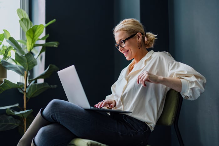 An investor smiles while sitting in a chair and looking at a laptop computer.