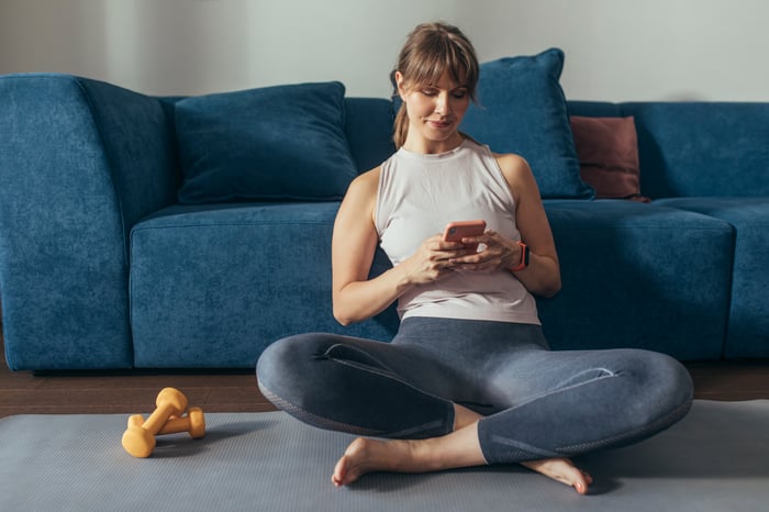 A woman, wearing yoga pants, sits atop a yoga mat while using her smartphone. A pair of weights lie next to her.